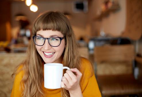 Smiling young adult woman wearing glasses and sipping coffee for a story on the best glasses for a heart shaped face