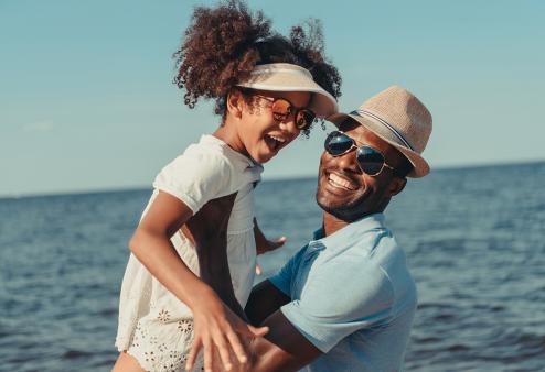 A father holding up his daughter at the beach, both wearing sunglasses